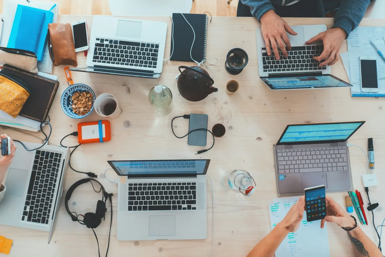 A busy collaborative workspace with multiple laptops, notebooks, smartphones, and other office essentials on a wooden table