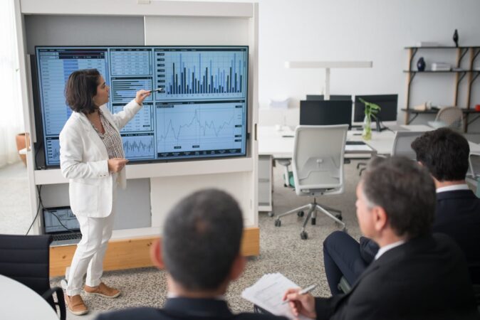 A woman in a white suit explaining data on a monitor to her colleagues